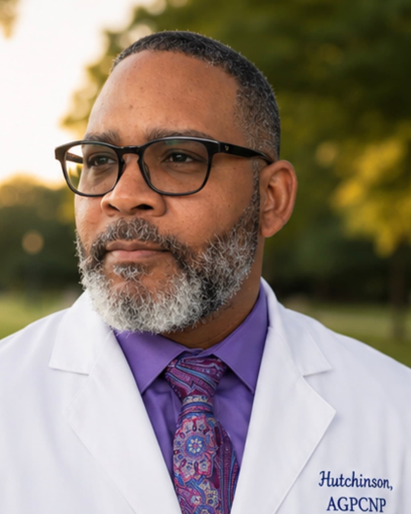 Al Hutchinson outdoors at golden hour in a white coat embroidered “Hutchinson, AGPCNP”, over a purple shirt and paisley tie.