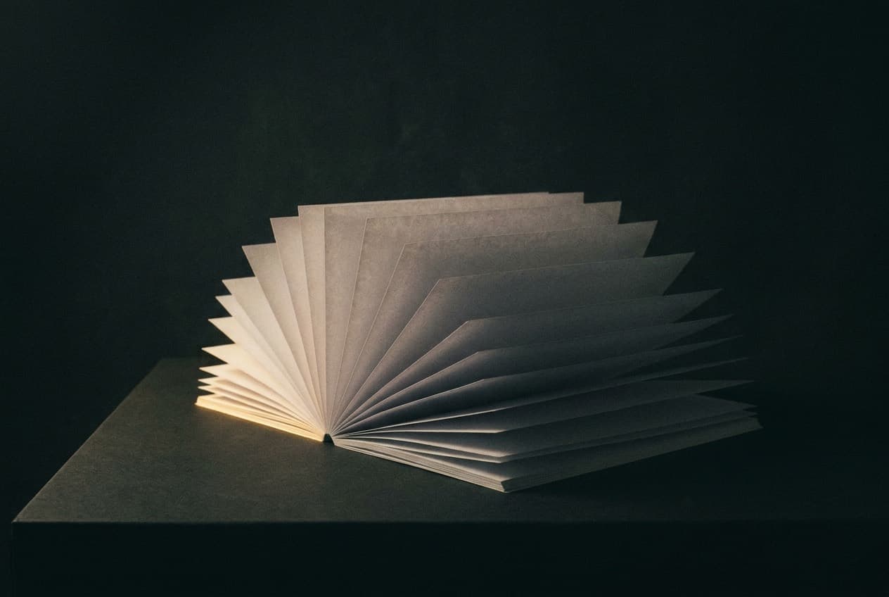 Sheets of blank paper fanned open in an otherwise dark room, their edges catching a single narrow warm light.